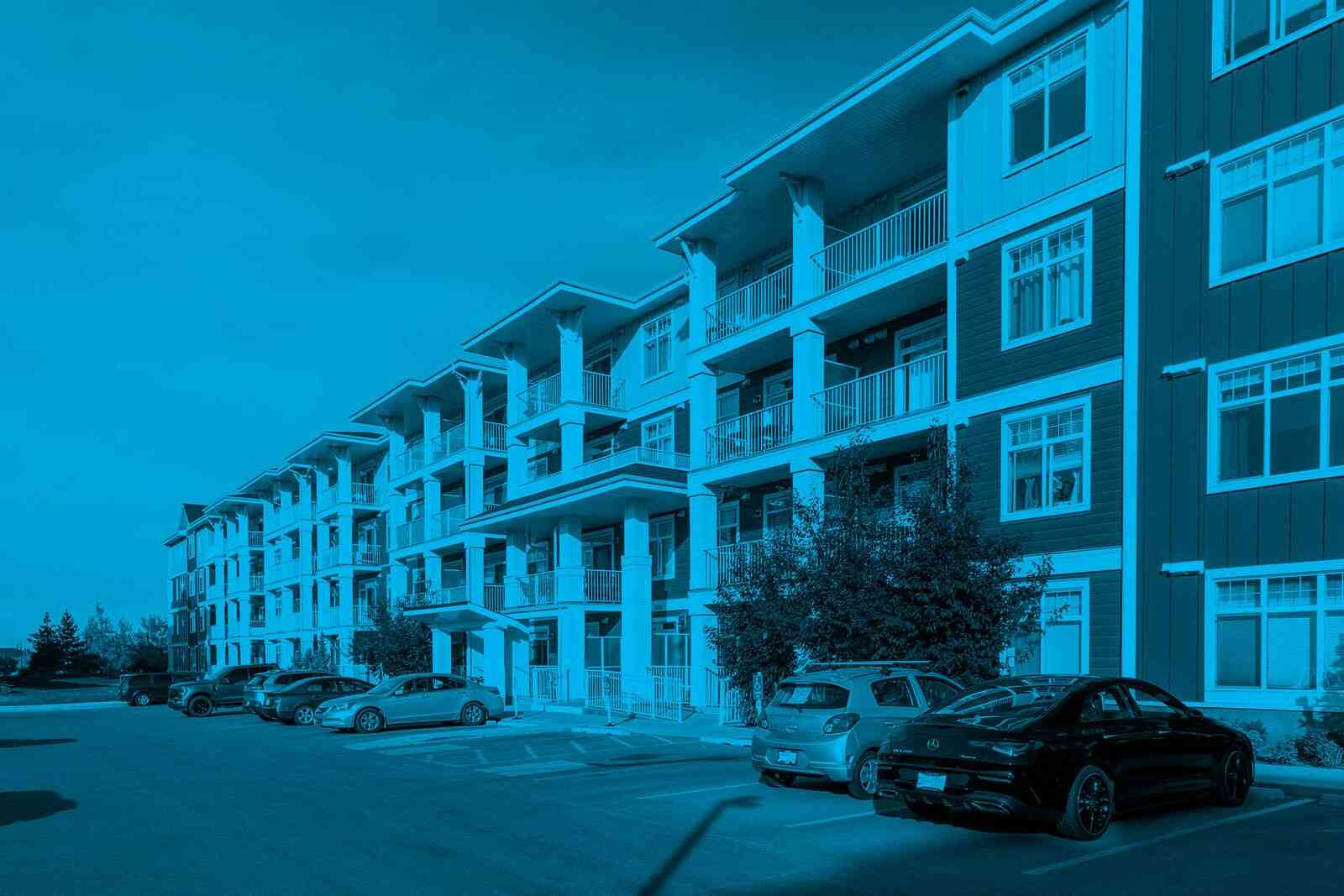 A modern multi-story condominium building featuring rows of private balconies and a paved parking lot with several cars parked in front, illustrating effective condo parking management.